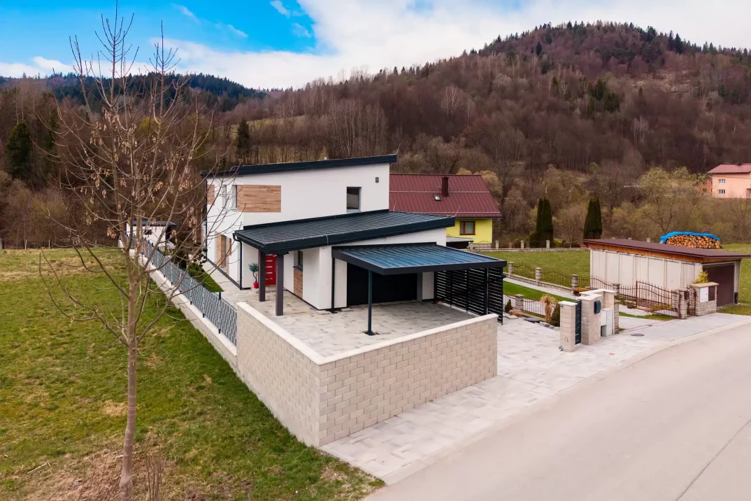 Modern családi házhoz épített Gardeon kocsibeálló, sík sötét tetővel és karcsú acél tartóoszloppal, panorámás hegyi kilátással