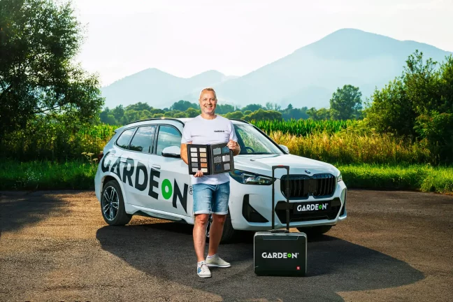Mosolygó GARDEON ügynök felületmintakönyvvel áll a cég BMW-je mellett.
