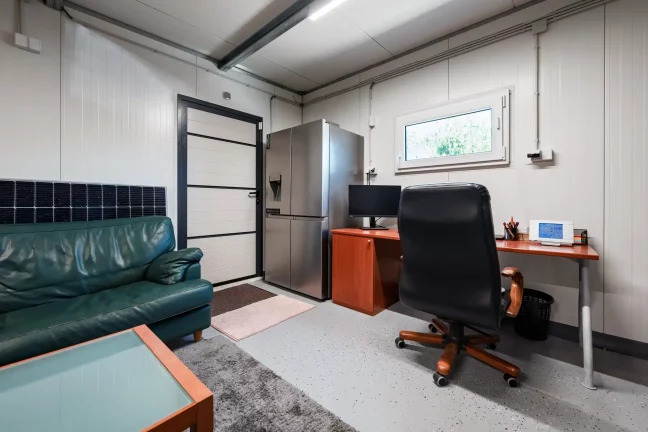 Helyiség zöld kanapéval, hűtőszekrénnyel és monitoros íróasztallal