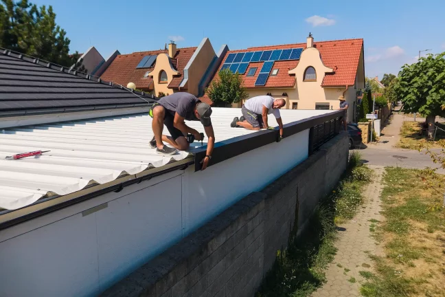 Két munkatárs az előtető tetején térdelve rögzíti a szegélylécet