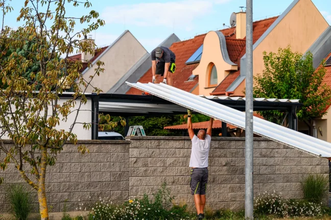 Egy munkatárs fehér tetőpanelt ad fel az előtető tetején dolgozó kollégájának