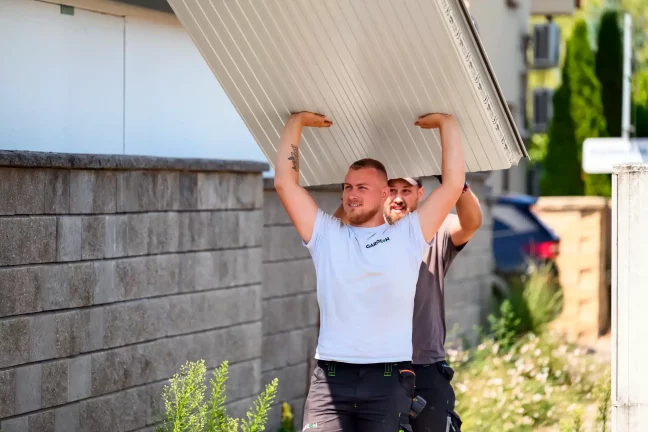 Két munkatárs egy nagy krémszínű tetőpanelt visz a feje felett