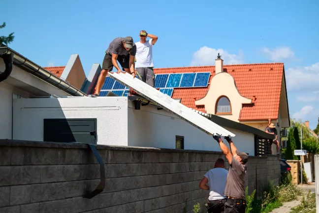 A tetőn dolgozó szerelők fehér panelt emelnek fel, amelyet egy kolléga alulról ad fel