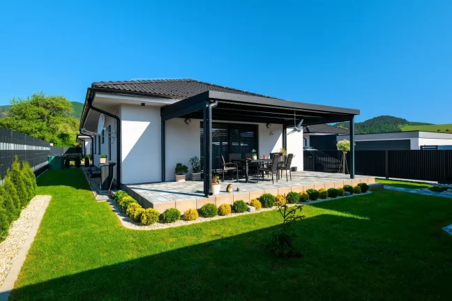 Pergola antracit színű szerkezettel egy fehér ház mellett, zöld gyeppel