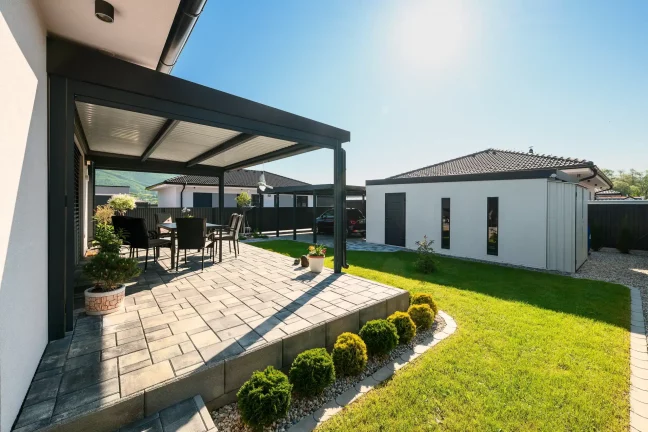 Egy antracit színű pergola oldalnézete teraszbútorokkal és gondozott kerttel a háznál