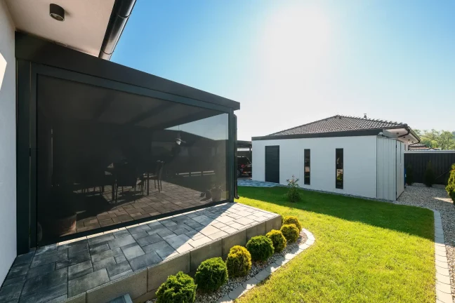 Antracit színű pergola leengedett rolóval egy fehér ház mellett, burkolt terasszal és zöld gyeppel