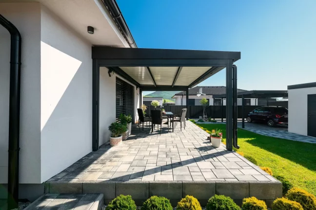 Antracit színű pergola lamellás tetővel egy fehér ház mellett, burkolt terasszal és kerti bútorokkal