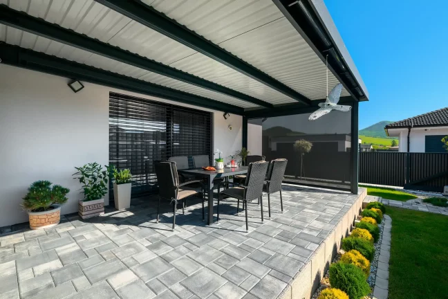 Kilátás a pergola belsejéből kerti bútorokra és teraszburkolatra, antracit színű szerkezet rolókkal