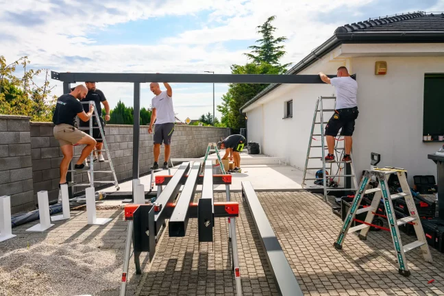 A szerelők létrák segítségével acélgerendát emelnek a ház fala mellett