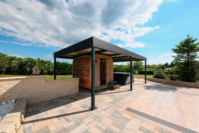 Szabadon álló antracit színű pergola lapos tetővel egy burkolt teraszon, háttérben zöld kerttel