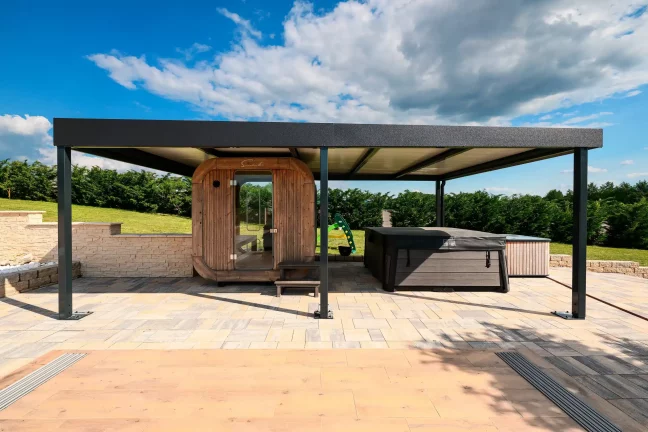 Antracit színű pergola lapos tetővel egy hordószauna mellett egy burkolt teraszon, kék éggel