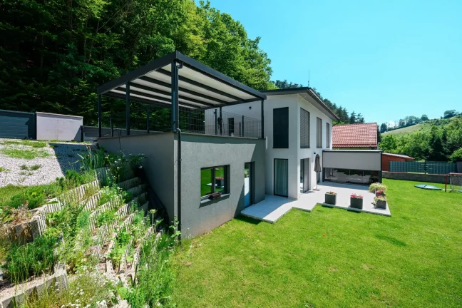 Pergola antracit acélszerkezettel és lapostetővel családi ház mellett.