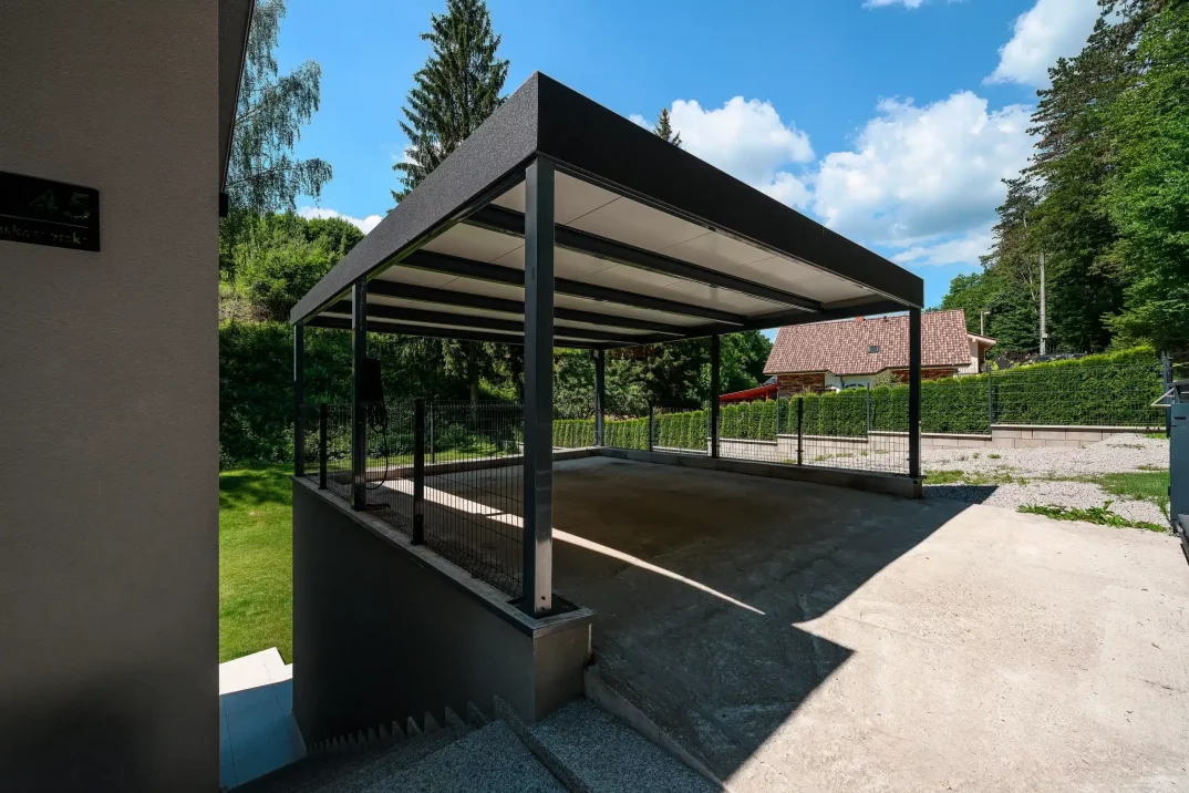 Sötétszürke kocsibeálló attikával végződő lapostetővel, családi ház mellett.