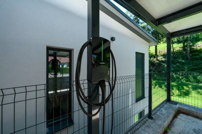 Elektromos autó töltője az antracit kocsibeálló oszlopán vakolt homlokzat mellett.