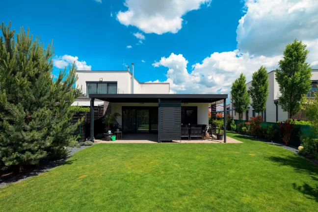 Antracit színű pergola lapos tetővel egy fehér ház mellett, tágas zöld gyeppel és fákkal