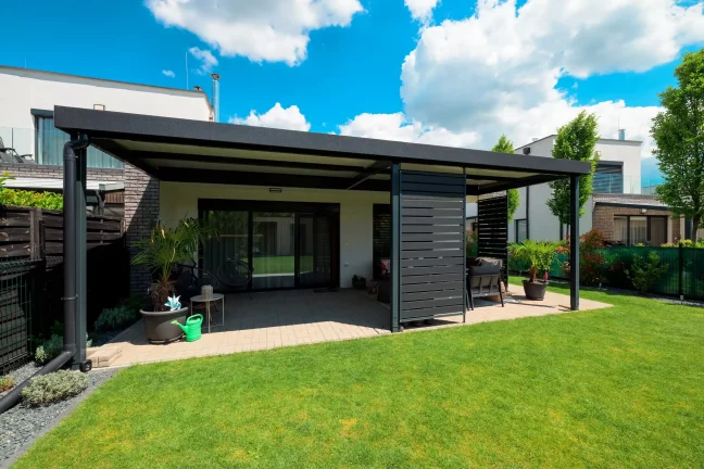 Szabadon álló antracit színű pergola lapos tetővel a zöld kertben, kék éggel és felhőkkel