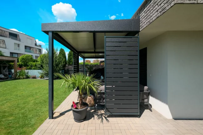 Antracit színű pergola oldalburkolattal egy téglaház mellett, dekoratív növényekkel a teraszon
